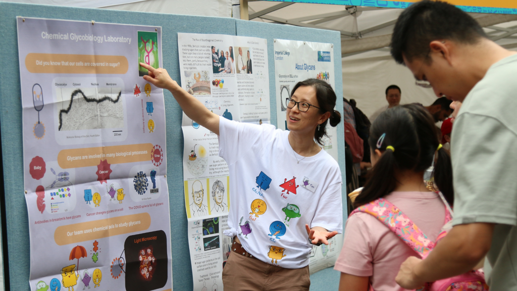 A female scientist wearing a t-shirt with Mr Men style illustrations of molecules points to a poster about Glycobiology. She is explaining her research to a father and daughter.